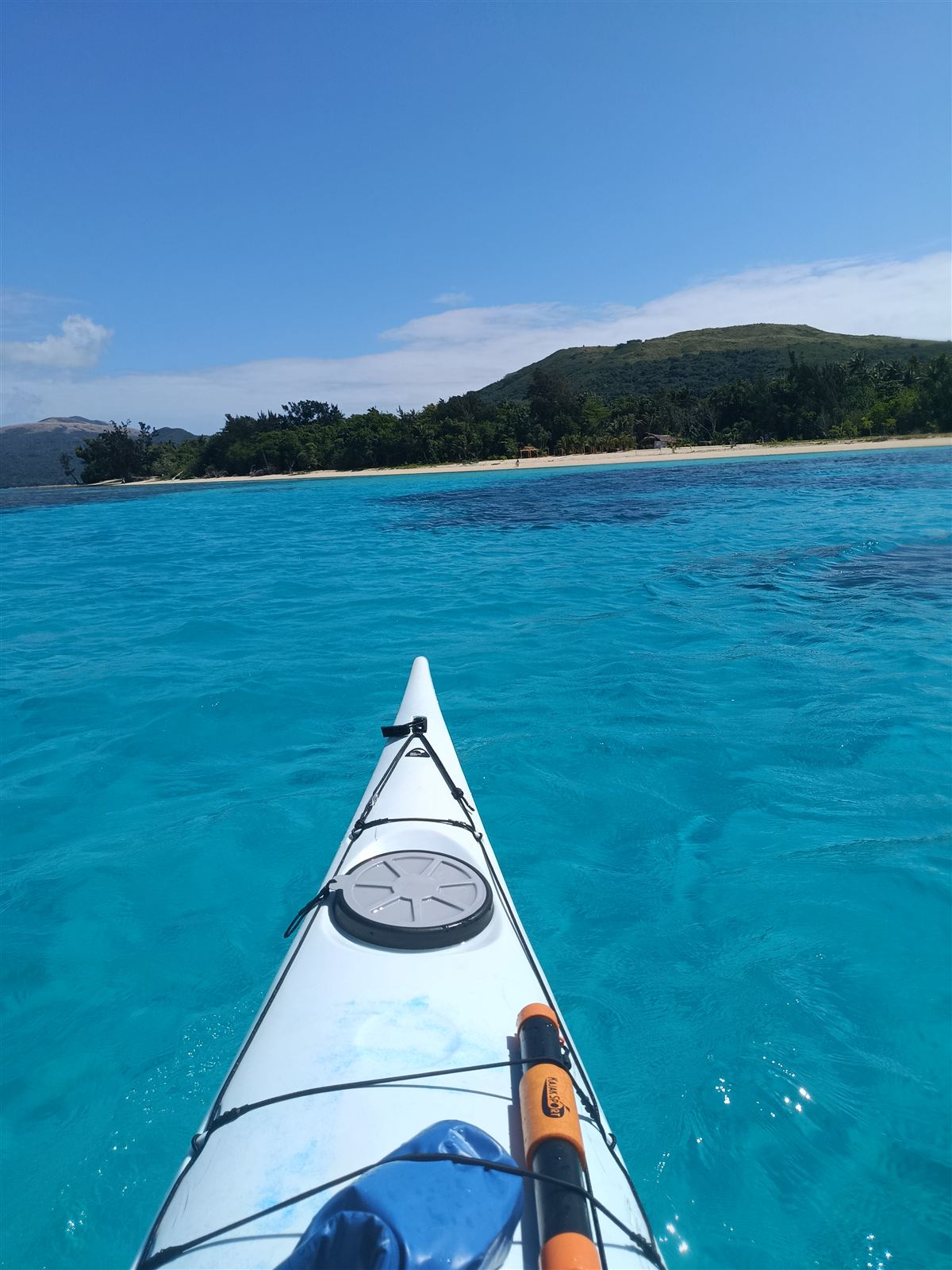 Kayak bow pointing across bright turquoise water toward a white sand beach and green hill